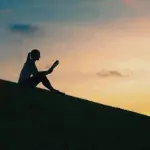 Sillhouette of seated woman reading a book with a sunset in the background.