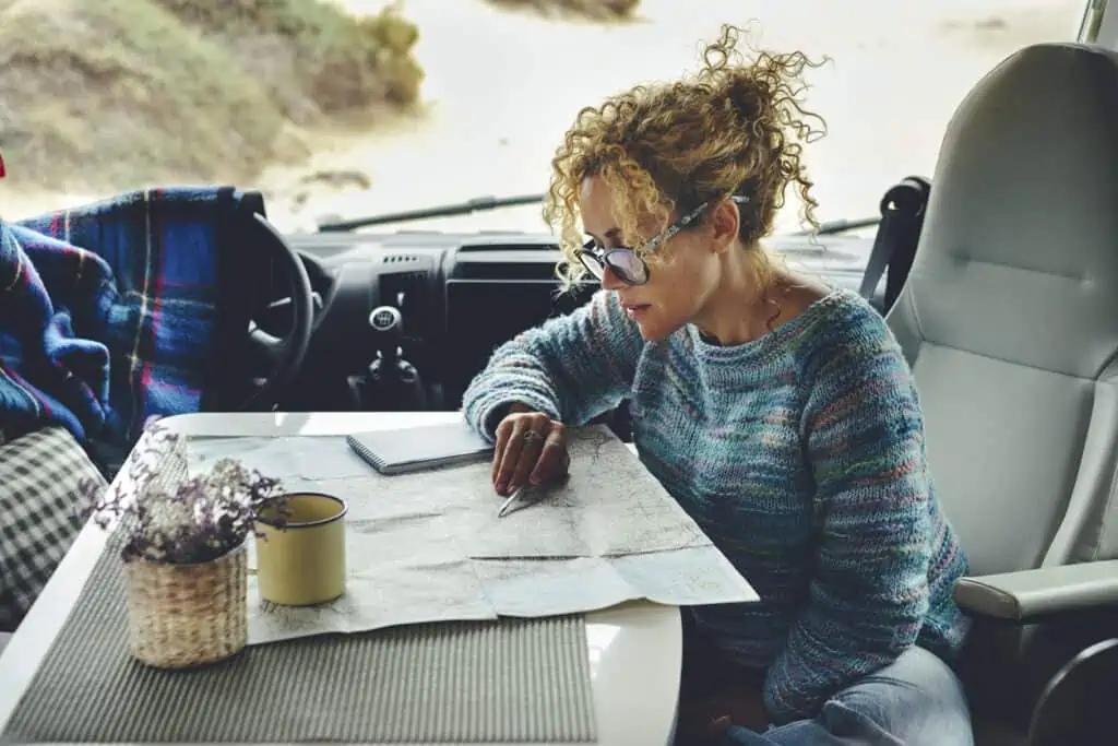 Adult woman traveling alone in her campervan sitting at table planning her road trip with a map and cellphone.